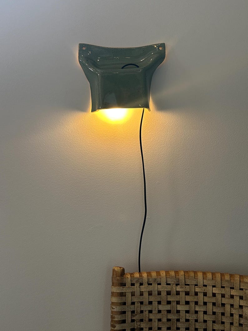 May include: A sage green ceramic wall sconce with a flared, sculptural shape. The fixture emits a warm, downward-facing glow and features a thin black power cord extending from the base. The light is mounted on a plain wall above a woven rattan surface.