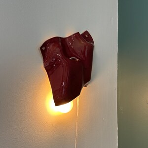 Puede incluir: Un aplique de pared de color rojo intenso y acabado brillante con una forma irregular y ondulada. La pieza presenta una abertura circular y emite una luz c&aacute;lida y brillante desde una bombilla situada en la parte inferior, con un cable transparente.