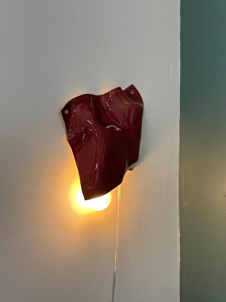 Puede incluir: Un aplique de pared de color rojo intenso y acabado brillante con una forma irregular y ondulada. La pieza presenta una abertura circular y emite una luz c&aacute;lida y brillante desde una bombilla situada en la parte inferior, con un cable transparente.