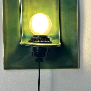 May include: A wall-mounted light fixture featuring a rectangular, glossy green ceramic backplate with a curved shelf. A black socket holds a round, frosted white light bulb that emits a warm glow. A black power cord extends downward from the base.