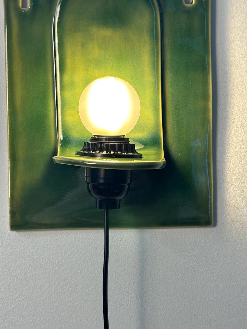 May include: A wall-mounted light fixture featuring a rectangular, glossy green ceramic backplate with a curved shelf. A black socket holds a round, frosted white light bulb that emits a warm glow. A black power cord extends downward from the base.