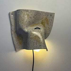 May include: A wall-mounted ceramic sconce with a speckled beige and tan finish. The fixture features an irregular, folded shape with two small circular holes at the top corners for mounting and a black power cord extending from the bottom light.