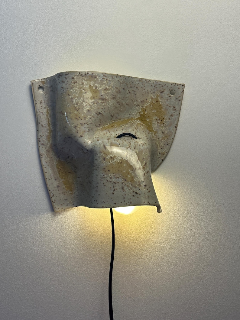 May include: A wall-mounted ceramic sconce with a speckled beige and tan finish. The fixture features an irregular, folded shape with two small circular holes at the top corners for mounting and a black power cord extending from the bottom light.