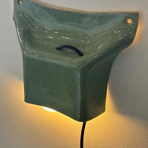 May include: A wall-mounted ceramic light fixture in a speckled sage green glaze. The piece features a flared, organic shape with two circular mounting holes at the top corners, a small black handle on the front, and a black power cord extending downward.