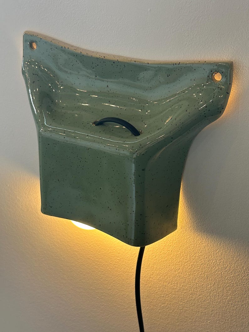 May include: A wall-mounted ceramic light fixture in a speckled sage green glaze. The piece features a flared, organic shape with two circular mounting holes at the top corners, a small black handle on the front, and a black power cord extending downward.