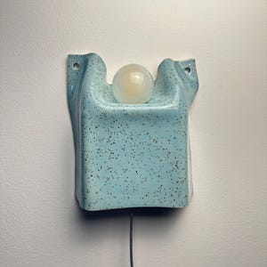 May include: A wall-mounted light fixture featuring a light blue ceramic base with a speckled brown pattern. The base has a curved, folded shape with two mounting holes at the top corners and a central circular opening holding a white glass globe bulb.