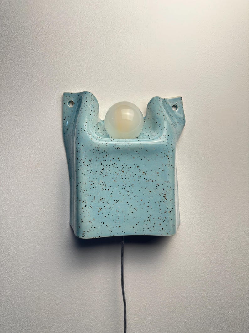 May include: A wall-mounted light fixture featuring a light blue ceramic base with a speckled brown pattern. The base has a curved, folded shape with two mounting holes at the top corners and a central circular opening holding a white glass globe bulb.