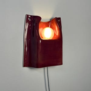 Puede incluir: Un aplique de pared de cer&aacute;mica en color burdeos intenso con acabado brillante y forma org&aacute;nica curva. La pieza presenta un recorte central que alberga una bombilla esf&eacute;rica brillante, con un cable de alimentaci&oacute;n fino en la base.