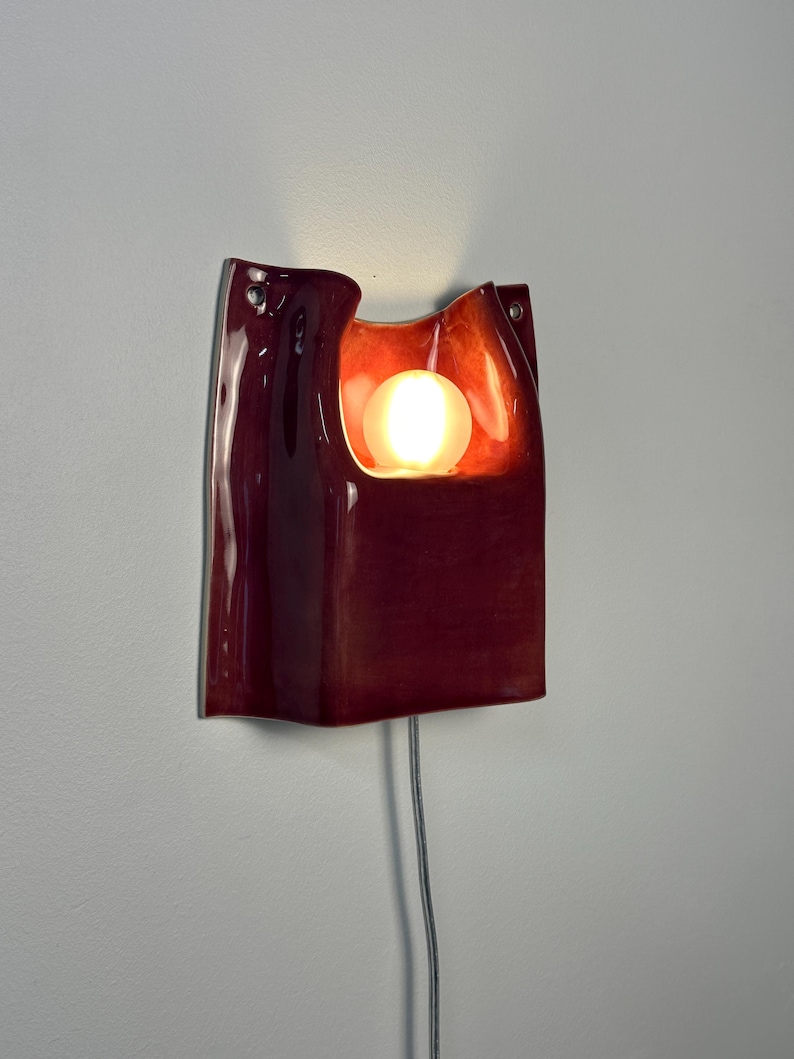 Puede incluir: Un aplique de pared de cer&aacute;mica en color burdeos intenso con acabado brillante y forma org&aacute;nica curva. La pieza presenta un recorte central que alberga una bombilla esf&eacute;rica brillante, con un cable de alimentaci&oacute;n fino en la base.