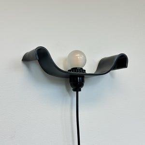 May include: A modern black wall sconce with a curved design. The sconce features a light bulb and a black cord. The fixture is mounted on a white wall.