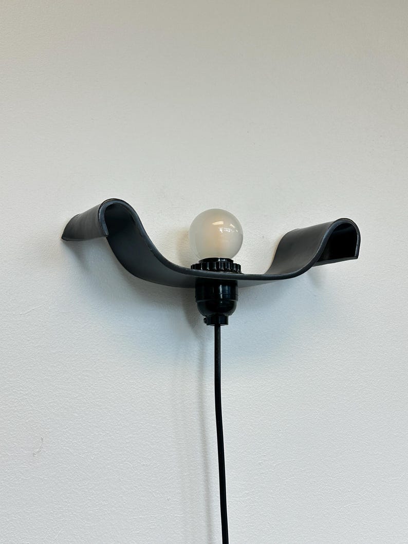 May include: A modern black wall sconce with a curved design. The sconce features a light bulb and a black cord. The fixture is mounted on a white wall.