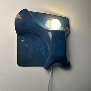 May include: A wall-mounted lamp featuring a glossy, deep blue ceramic base with an irregular, organic shape. A single, spherical white light bulb is nestled within a recessed section of the blue ceramic, emitting a warm glow downward.