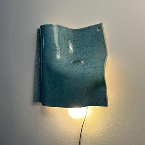 May include: A wall-mounted light fixture featuring a curved, speckled light blue ceramic shade. The shade has a glossy finish with dark flecks and a small circular opening near the top. A single warm-toned light bulb hangs below the ceramic piece.