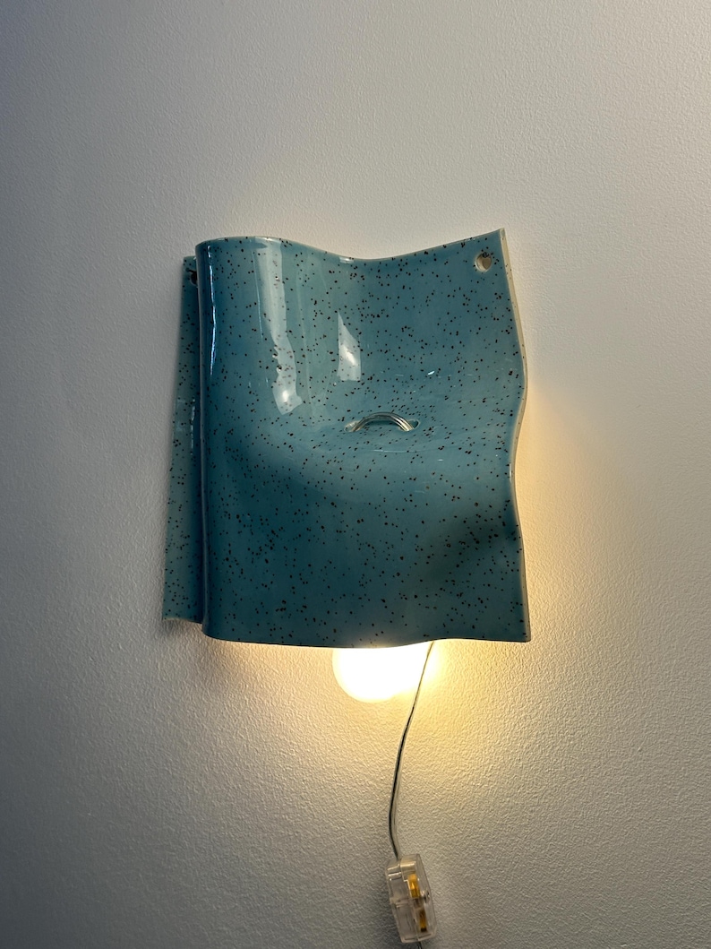 May include: A wall-mounted light fixture featuring a curved, speckled light blue ceramic shade. The shade has a glossy finish with dark flecks and a small circular opening near the top. A single warm-toned light bulb hangs below the ceramic piece.