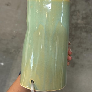 May include: A cylindrical ceramic lamp base with a glossy, sage green glaze featuring subtle tonal variations. The surface is smooth with a slightly irregular, organic shape. A clear electrical cord exits through a circular hole near the base.
