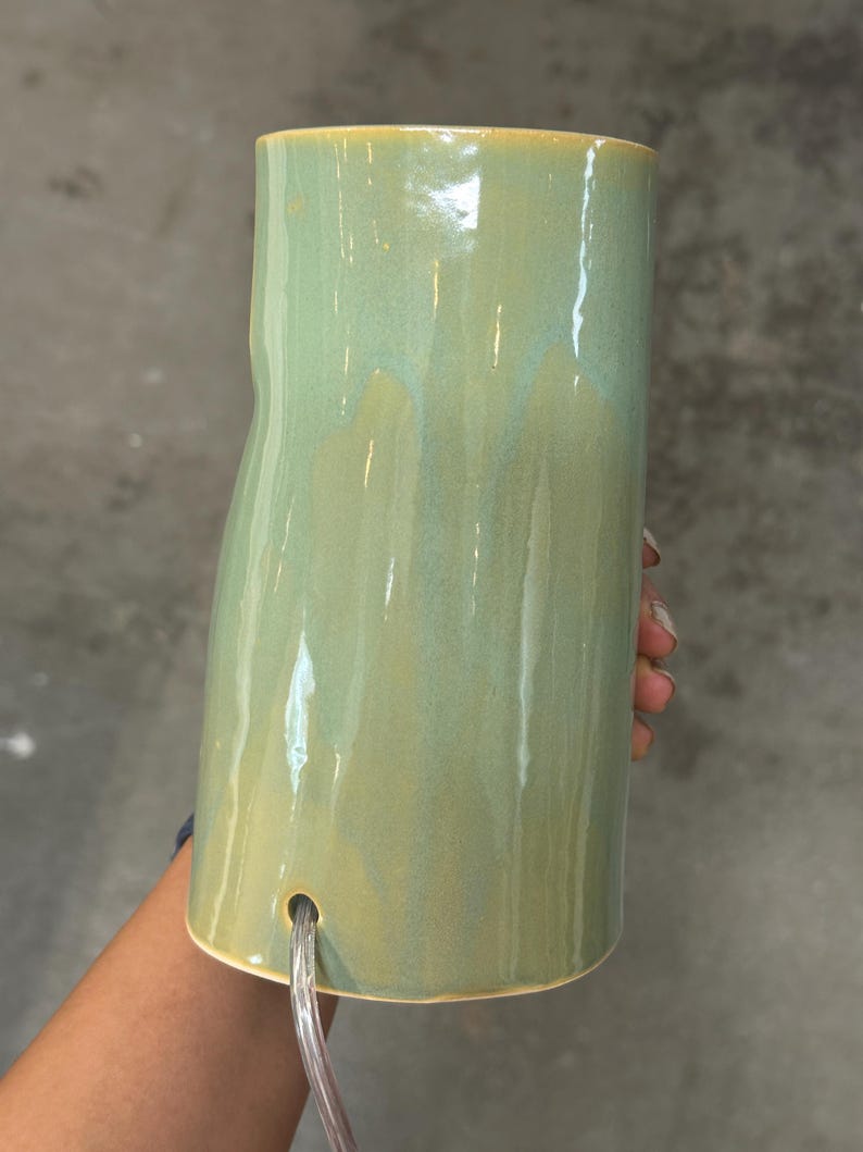 May include: A cylindrical ceramic lamp base with a glossy, sage green glaze featuring subtle tonal variations. The surface is smooth with a slightly irregular, organic shape. A clear electrical cord exits through a circular hole near the base.
