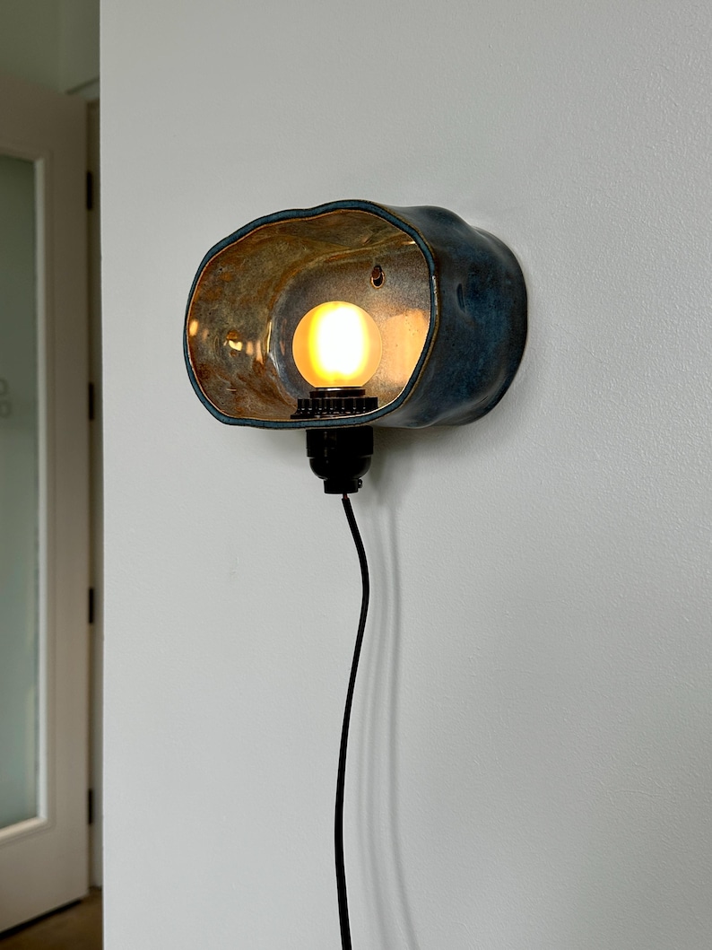 May include: A wall-mounted ceramic light fixture with a blue and brown glazed finish. The fixture has an organic shape, with a warm light bulb inside. A black cord extends downward.