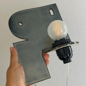 May include: A wall-mounted ceramic light fitting featuring a smooth, slate-grey, irregular geometric backplate with two circular mounting holes. A black socket holds a round, frosted white light bulb, connected by a clear electrical cord.