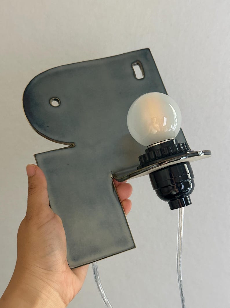 May include: A wall-mounted ceramic light fitting featuring a smooth, slate-grey, irregular geometric backplate with two circular mounting holes. A black socket holds a round, frosted white light bulb, connected by a clear electrical cord.