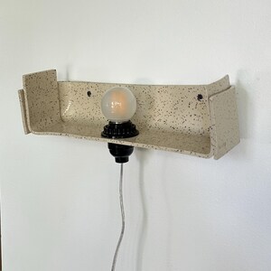 May include: A speckled beige ceramic wall sconce with a black bulb socket and a round, frosted light bulb. The sconce has a shelf-like design with a curved front and two side panels. A silver cord hangs down.