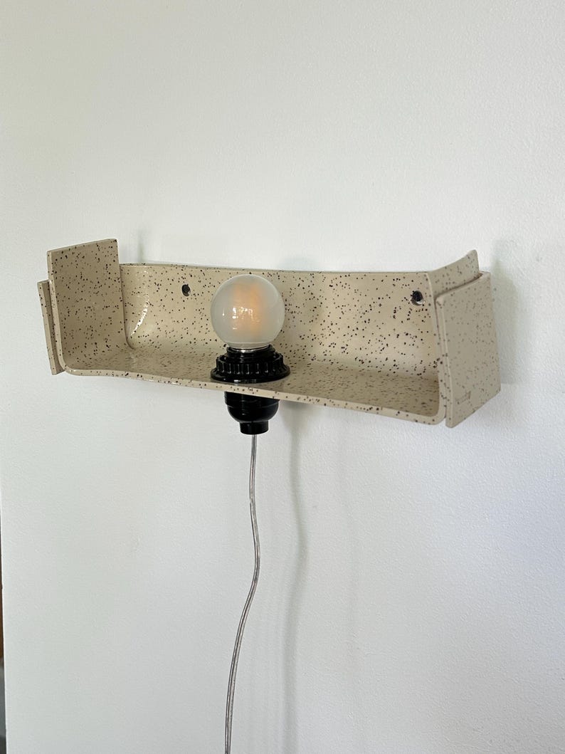 May include: A speckled beige ceramic wall sconce with a black bulb socket and a round, frosted light bulb. The sconce has a shelf-like design with a curved front and two side panels. A silver cord hangs down.