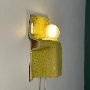 Puede incluir: Un aplique de pared con una base de cer&aacute;mica amarilla curvada con un patr&oacute;n de motas negras. La l&aacute;mpara incluye una bombilla esf&eacute;rica de vidrio esmerilado que emite un brillo c&aacute;lido y suave, con un cable de alimentaci&oacute;n transparente.