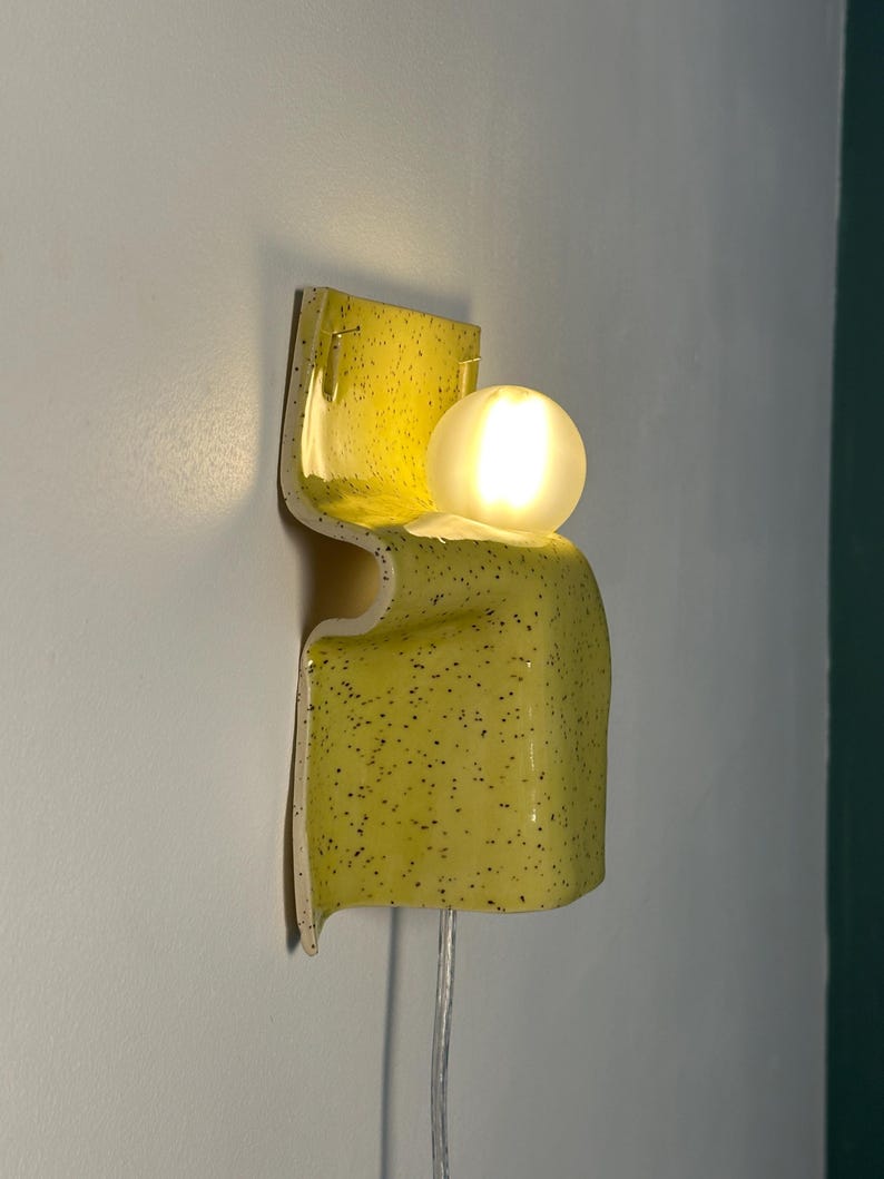 Puede incluir: Un aplique de pared con una base de cer&aacute;mica amarilla curvada con un patr&oacute;n de motas negras. La l&aacute;mpara incluye una bombilla esf&eacute;rica de vidrio esmerilado que emite un brillo c&aacute;lido y suave, con un cable de alimentaci&oacute;n transparente.