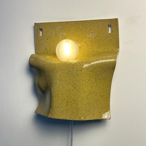 May include: A wall-mounted ceramic sconce featuring a speckled mustard yellow glaze with a curved, folded form. A single spherical frosted glass light bulb sits in the center of the fixture, emitting a warm glow. A thin power cord hangs below.