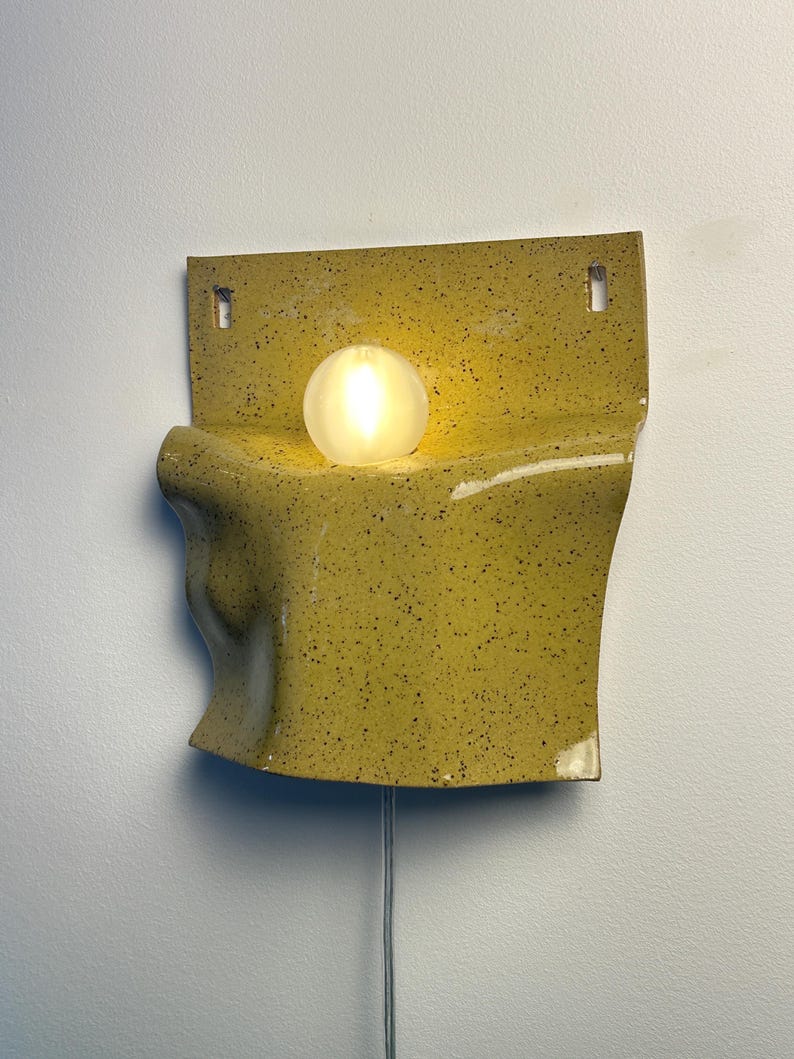May include: A wall-mounted ceramic sconce featuring a speckled mustard yellow glaze with a curved, folded form. A single spherical frosted glass light bulb sits in the center of the fixture, emitting a warm glow. A thin power cord hangs below.