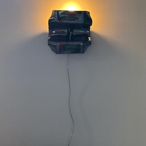 May include: A dark, glossy, rectangular wall sconce with a curved, molded form. The fixture emits a warm, golden light from its top opening and features a thin, light-colored electrical cord extending downward from the base of the unit.