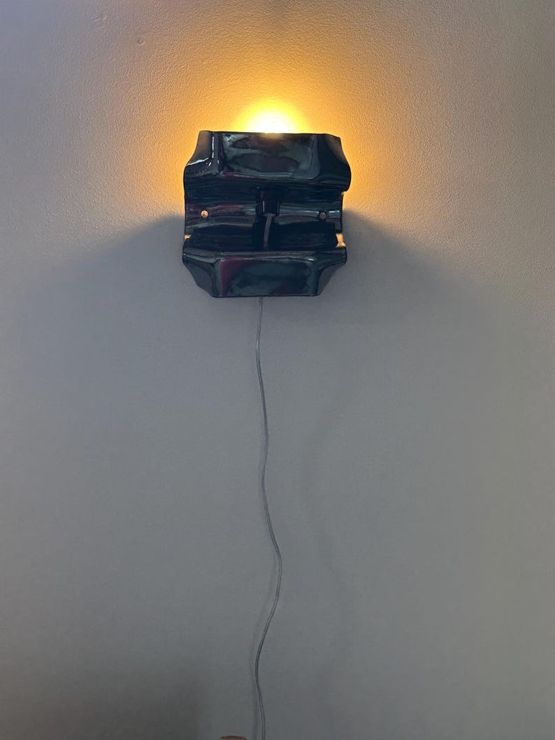 May include: A dark, glossy, rectangular wall sconce with a curved, molded form. The fixture emits a warm, golden light from its top opening and features a thin, light-colored electrical cord extending downward from the base of the unit.