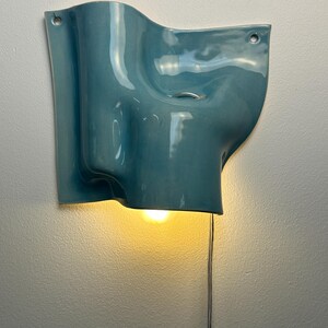 May include: A wall-mounted ceramic light fitting with a glossy, teal-blue finish and an organic, undulating shape. The piece features two circular mounting holes at the top corners and emits a warm, downward-facing glow from a single bulb.