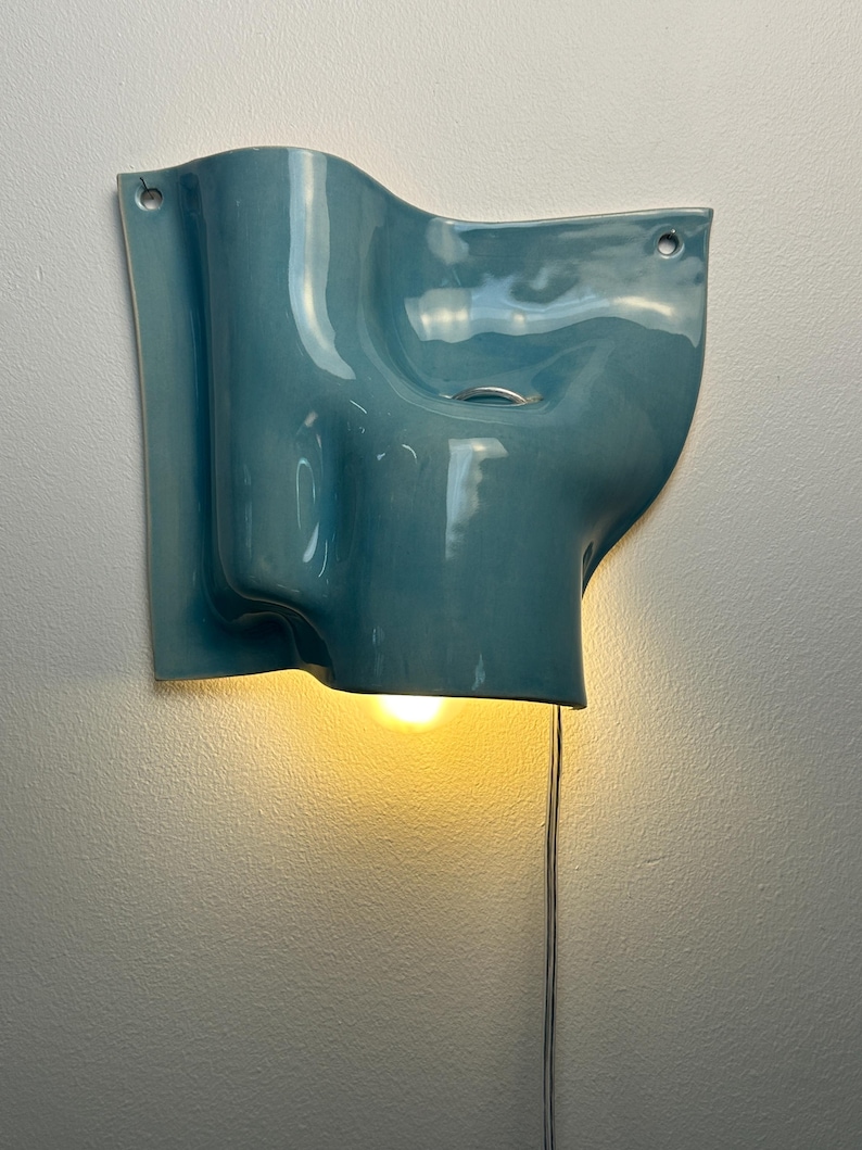 May include: A wall-mounted ceramic light fitting with a glossy, teal-blue finish and an organic, undulating shape. The piece features two circular mounting holes at the top corners and emits a warm, downward-facing glow from a single bulb.