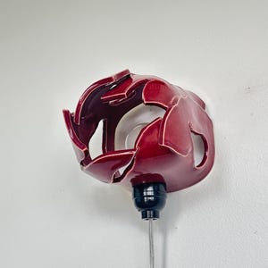 May include: A red ceramic wall sconce with a glossy finish. The sconce has an open design with petal-like shapes. It features a black base and a silver cord, mounted on a white wall.