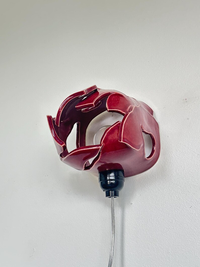 May include: A red ceramic wall sconce with a glossy finish. The sconce has an open design with petal-like shapes. It features a black base and a silver cord, mounted on a white wall.