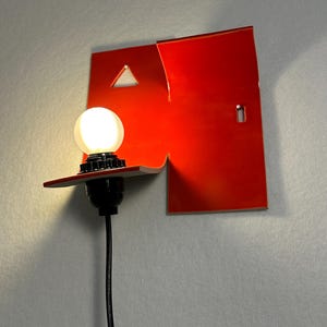 May include: A contemporary red wall lamp with a distinctive geometric shape. The lamp has a triangular cut-out and a small rectangular opening. A round light bulb is lit, connected to a black cable.