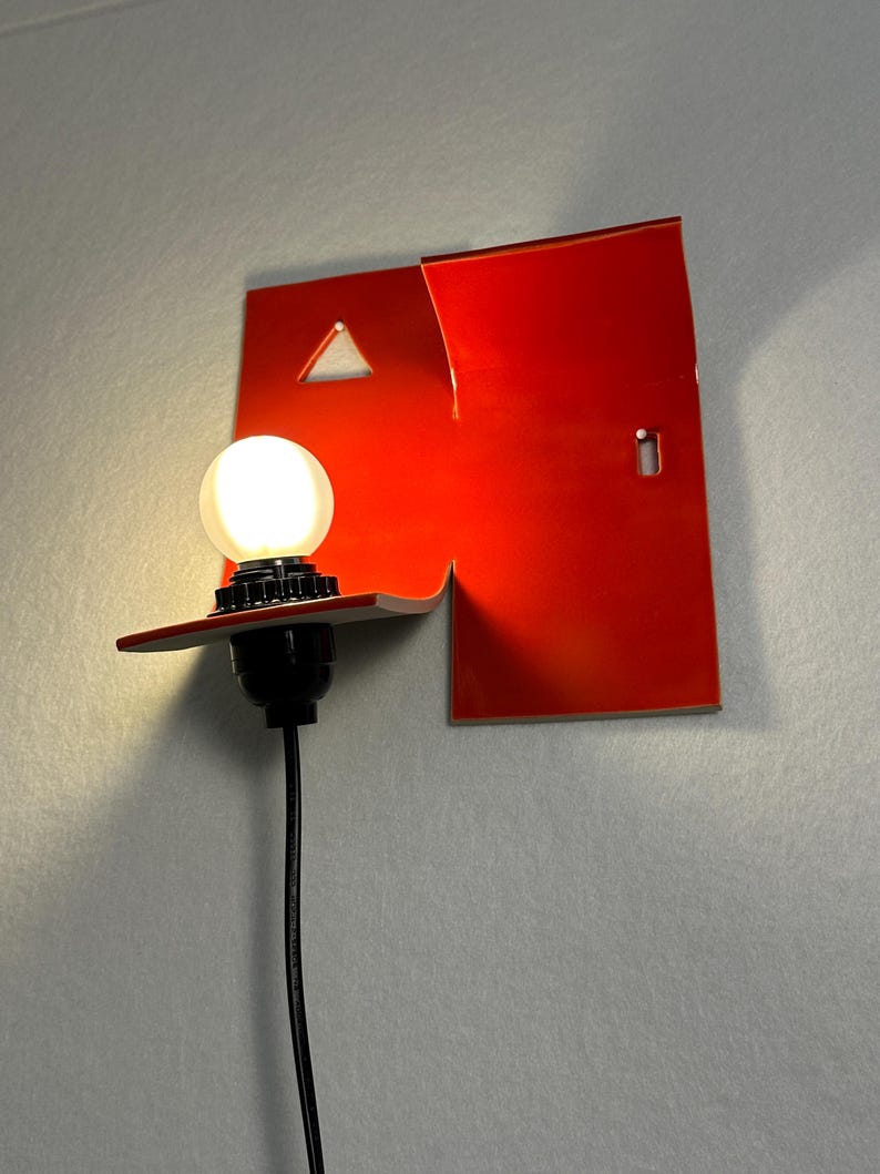 May include: A contemporary red wall lamp with a distinctive geometric shape. The lamp has a triangular cut-out and a small rectangular opening. A round light bulb is lit, connected to a black cable.
