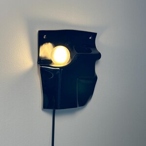 Puede incluir: Un aplique de pared negro con acabado brillante y forma moldeada irregular. La l&aacute;mpara cuenta con una bombilla redonda que emite una luz c&aacute;lida desde una abertura empotrada en el lado izquierdo, con un cable de alimentaci&oacute;n negro hacia abajo.