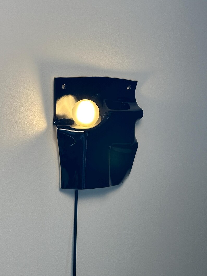 Puede incluir: Un aplique de pared negro con acabado brillante y forma moldeada irregular. La l&aacute;mpara cuenta con una bombilla redonda que emite una luz c&aacute;lida desde una abertura empotrada en el lado izquierdo, con un cable de alimentaci&oacute;n negro hacia abajo.