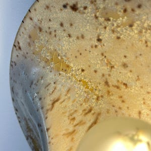 May include: A curved ceramic wall sconce featuring a glossy cream-coloured glaze with brown speckles and a distinct crystalline texture. The surface displays a crackled pattern with yellow and amber tones, illuminated by a round, warm-toned light bulb.
