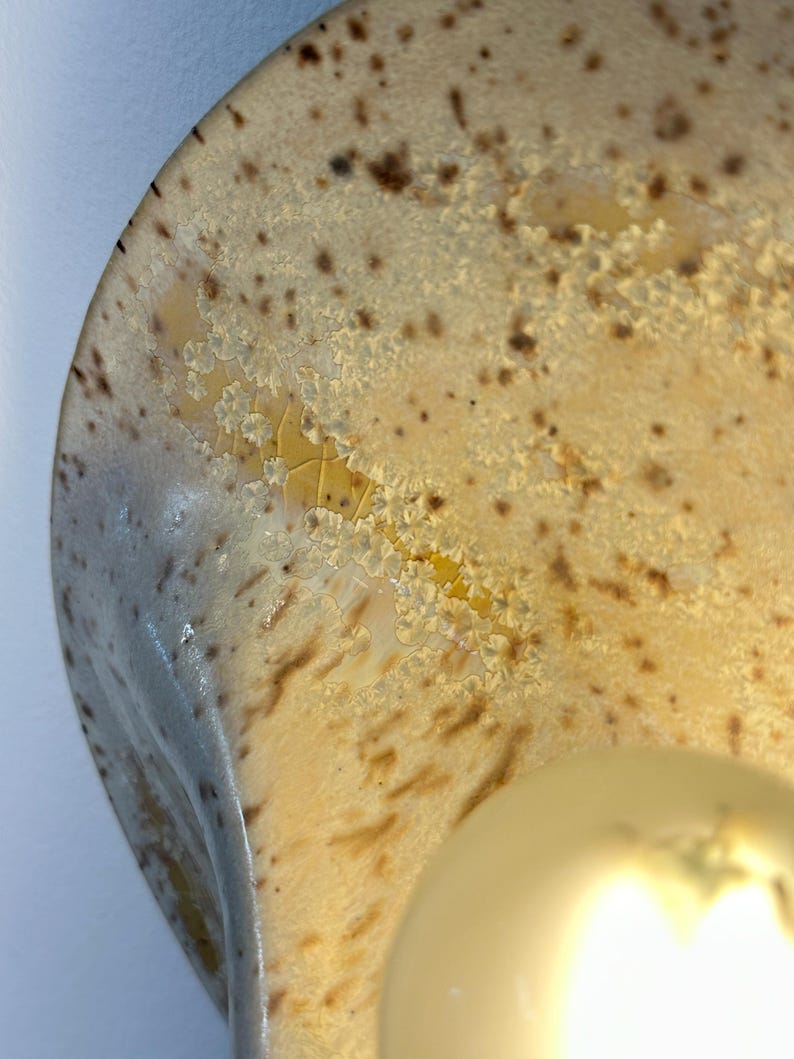 May include: A curved ceramic wall sconce featuring a glossy cream-coloured glaze with brown speckles and a distinct crystalline texture. The surface displays a crackled pattern with yellow and amber tones, illuminated by a round, warm-toned light bulb.