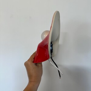 May include: A wall-mounted lamp featuring a glossy red curved shade with a white interior. The fixture includes a round light bulb, a black socket, and exposed black and white electrical wires extending from the base of the lamp housing.