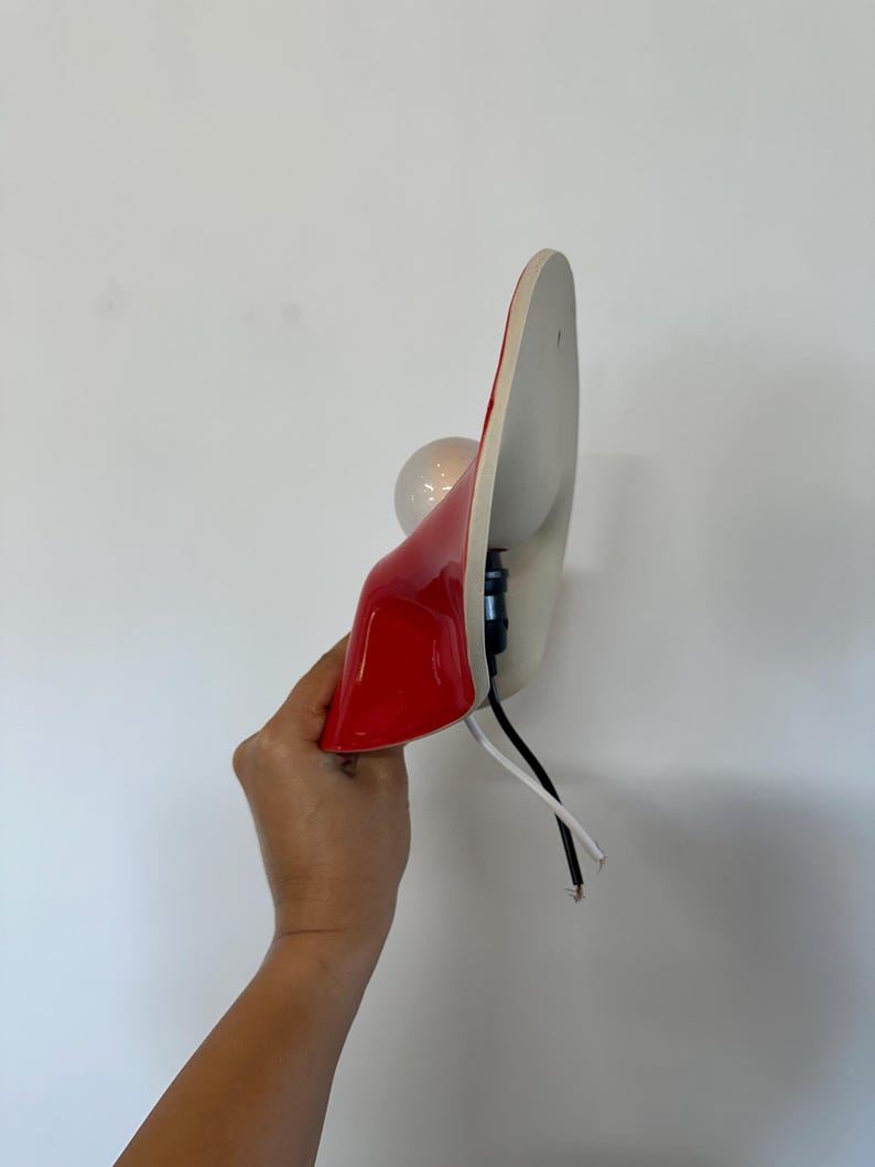 May include: A wall-mounted lamp featuring a glossy red curved shade with a white interior. The fixture includes a round light bulb, a black socket, and exposed black and white electrical wires extending from the base of the lamp housing.