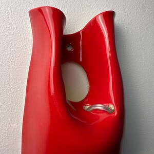 May include: A glossy red ceramic wall vase with a smooth, curved silhouette. The piece features a central cutout opening and a small hole near the top for mounting. A clear, flexible plastic tube is visible inside the lower section of the opening.