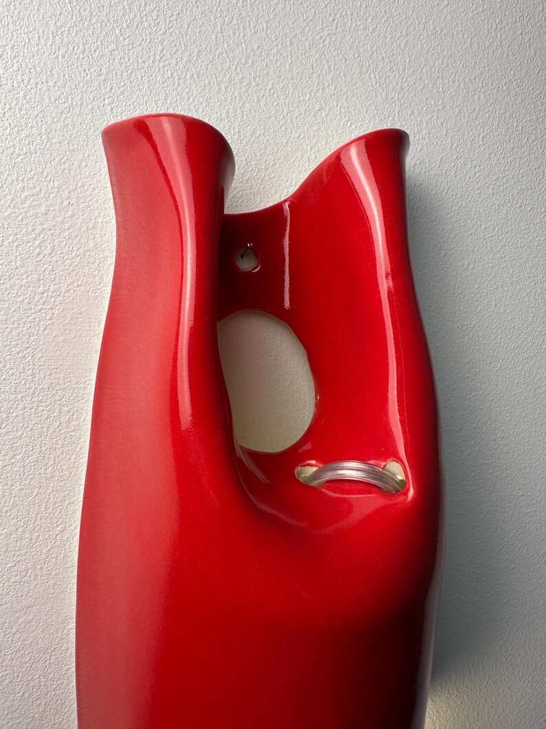 May include: A glossy red ceramic wall vase with a smooth, curved silhouette. The piece features a central cutout opening and a small hole near the top for mounting. A clear, flexible plastic tube is visible inside the lower section of the opening.