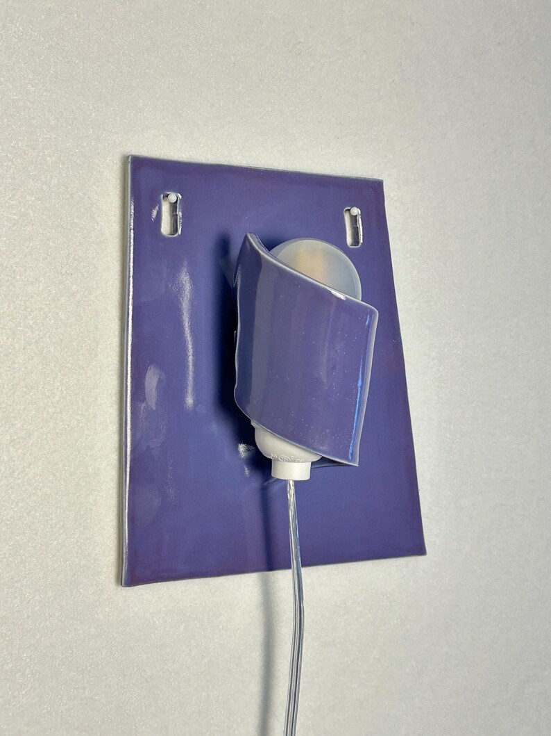 Pode incluir: Arandela de parede moderna com base de cer&acirc;mica roxa e abajur curvo na cor lavanda. A arandela apresenta uma l&acirc;mpada branca e um cabo de alimenta&ccedil;&atilde;o transparente. Design minimalista e contempor&acirc;neo, adequado para ilumina&ccedil;&atilde;o interior.