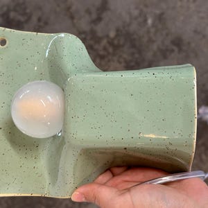May include: A wall-mounted ceramic light fitting in a light sage green colour with dark brown speckles. The fitting features a rectangular backplate with two circular mounting holes and a protruding, curved housing that holds a round white light bulb.
