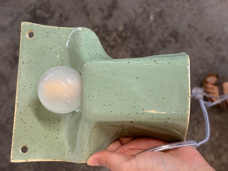 May include: A wall-mounted ceramic light fitting in a light sage green colour with dark brown speckles. The fitting features a rectangular backplate with two circular mounting holes and a protruding, curved housing that holds a round white light bulb.
