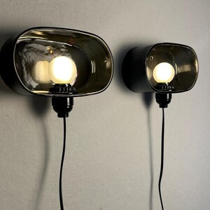 May include: Two black wall lamps with oval shades. Each lamp has a glowing light bulb and a black power cord. The shades have a reflective, metallic interior. The lamps are mounted on a light grey wall.