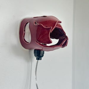 May include: A burgundy ceramic wall sconce with an unusual, organic form. It features a bulb socket and a silver cord. The sconce is mounted on a white wall.